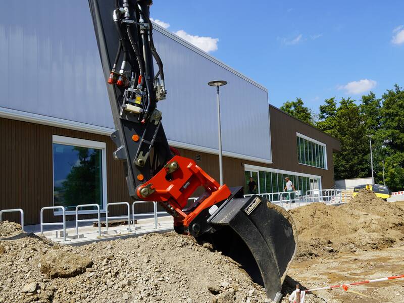 Ein Bagger hebt Erde auf einer Baustelle vor der Sporthalle 1 in Nellingen, die über große Fenster und hellbrauner Fassade verfügt, aus.
