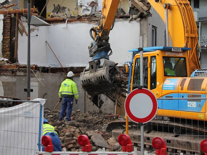 Eine Baustelle zeigt einen großen Bagger, der ein Gebäude abreißt, während Arbeiter in Schutzkleidung daneben stehen.