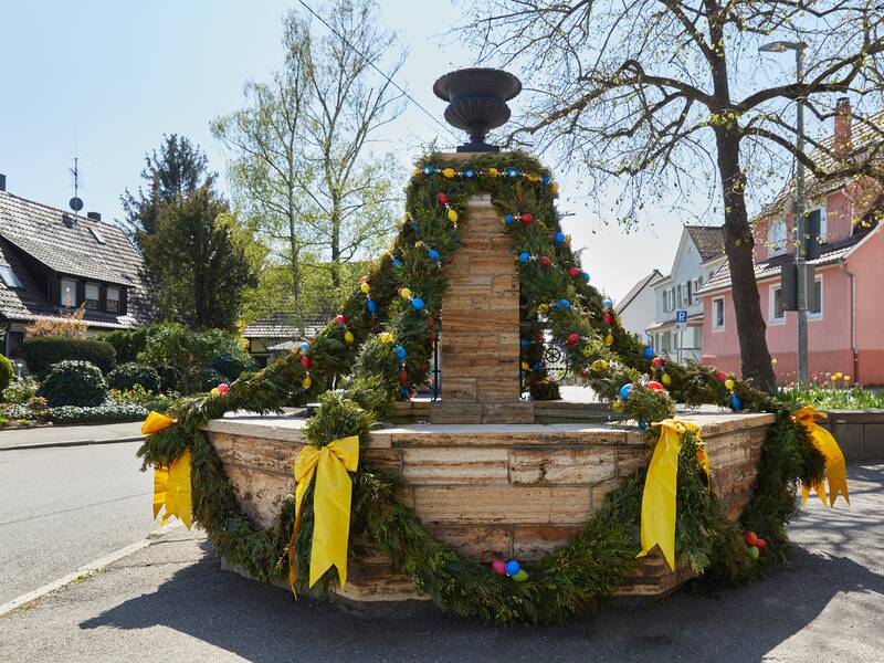 Der Kemnater Brunnen ist mit Tannenzweigen, bunten Ostereiern und gelben Schleifen als Osterbrunnen geschmückt. Er steht in einer Straße, umgeben von Häusern und Bäumen.