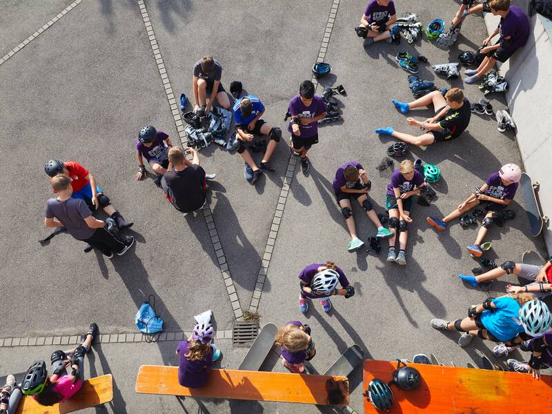 Eine Gruppe von Jugendlichen mit Helmen und Schutzkleidung zum Inline-Skaten oder Skateboard-Fahren sitzt auf dem Boden und macht Pause.