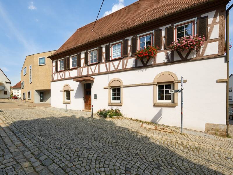 Das Bild zeigt eine gepflasterte Straße mit dem Alten Kemnater Rathaus, das mit Blumenkästen dekoriert ist, sowie modernen Gebäuden im Hintergrund.
