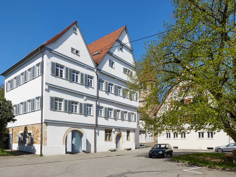 Das Bild zeigt das Nellinger Rathaus, ein großes, weißes, mehrstöckiges Gebäude mit rotem Dach und vielen Fenstern, umgeben von einem Baum und einem Parkplatz, bei klarem Wetter.