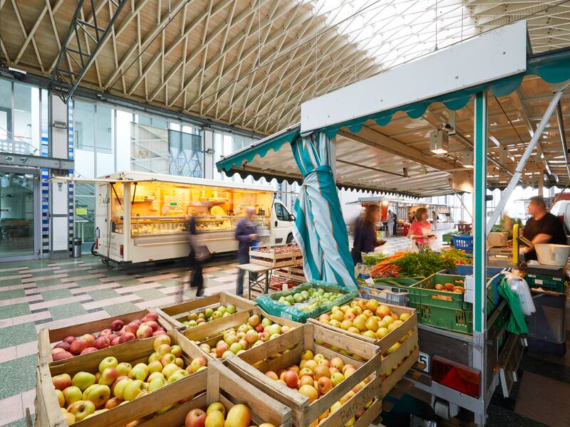 In Nellingen befindet sich An der Halle ein überdachter Wochenmarkt mit Verkaufsständen und Kisten voller Obst und Gemüse, an denen Menschen vorbeigehen.