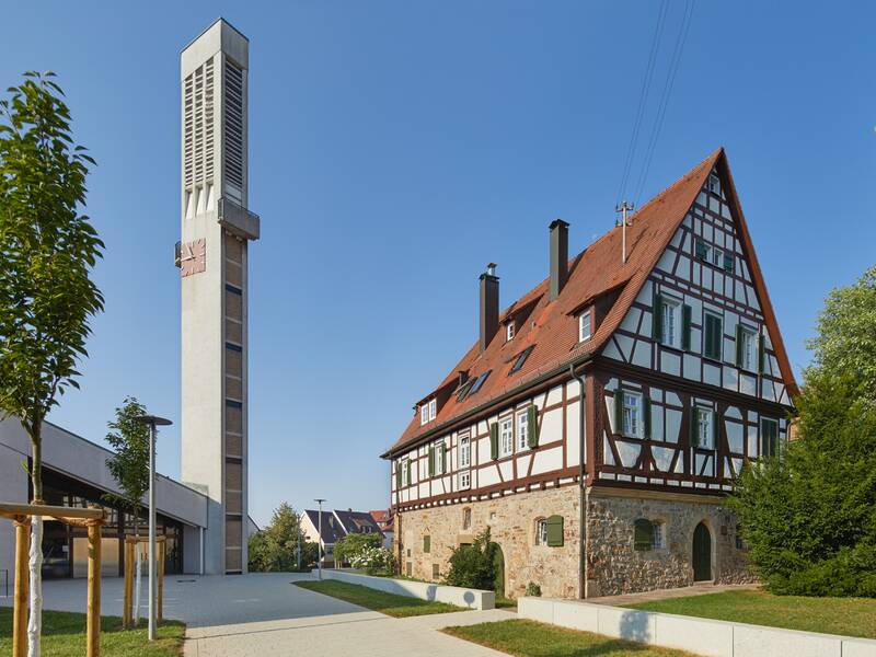 Das Bild zeigt eine Kirche mit modernem, schmalen Glockenturm neben einem traditionellen Fachwerkhaus, dem Pfarrhaus,welches rote Dachziegel und grünen Fensterläden hat und umgeben von Bäumen und einem Gehweg ist.