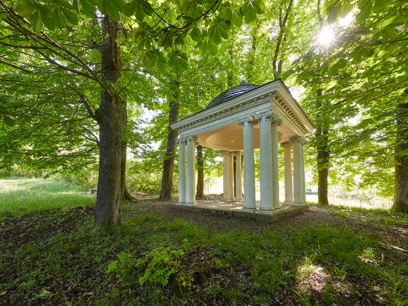 Ein kleiner, runder Pavillon mit Säulen, der sogenannte Amortempel, steht in einem von Sonnenlicht durchfluteten, grünen Waldstück.