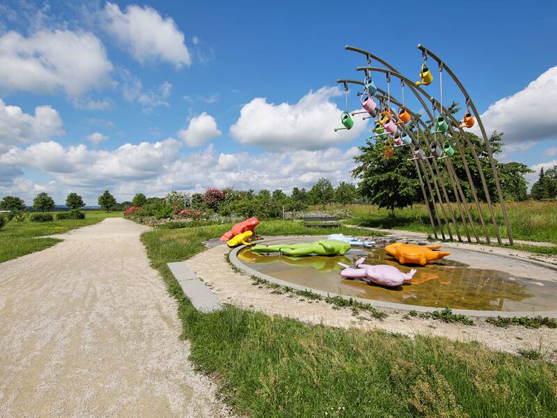 Der Froschbrunnen im Scharnhauser Park hat auf der Wasserfläche große, bunte Frösche, über denen an langen gebogenen Stangen ebenso bunte Gießkannen hängen.