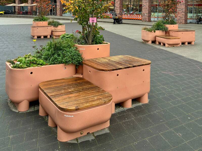 Auf einem gepflasterten Platz stehen mehrere modulare Sitzgelegenheiten aus orangefarbenem Material mit holzverkleideten Sitzen und integrierten Pflanzkästen, die mit verschiedenen Pflanzen gefüllt sind.