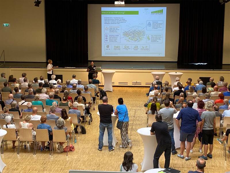 In einem Saal sitzen zahlreiche Menschen vor einer Bühne, auf der zwei Personen stehen und eine Präsentation mit einem großen Diagramm zeigen. (KI generiert)