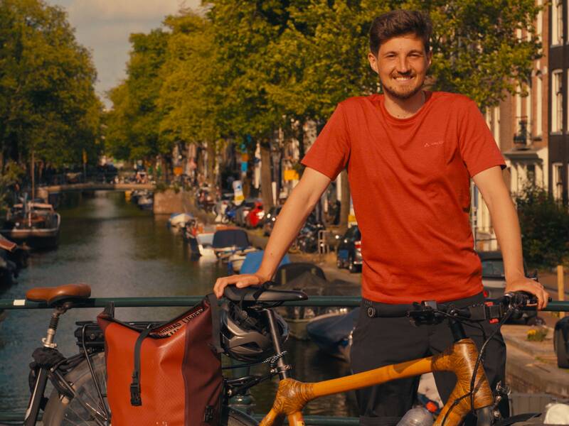 Ingwar Perowanowitsch steht lächelnd neben einem Fahrrad auf einer Brücke über einem von Bäumen gesäumten Kanal mit Booten.