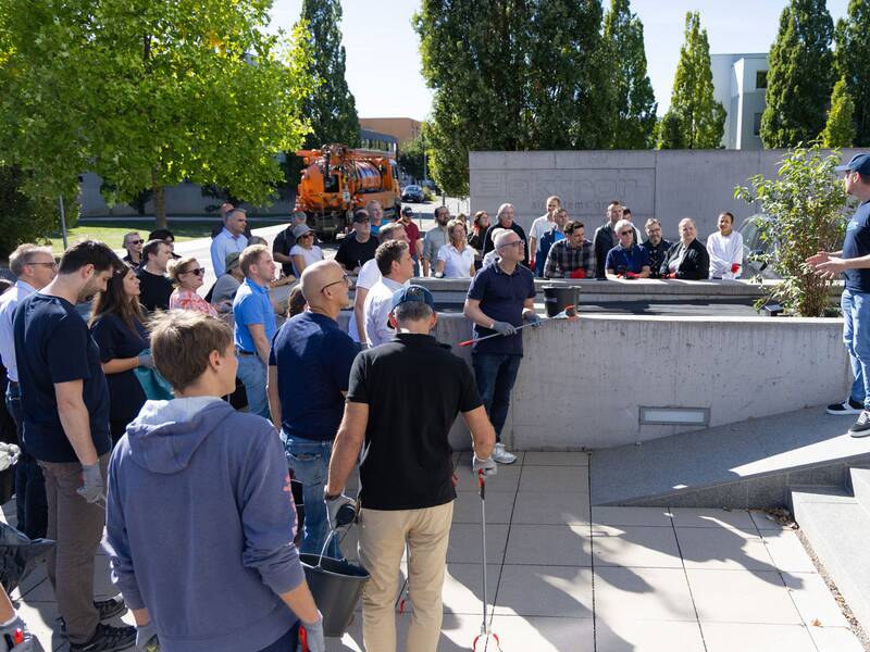 Eine Gruppe von Menschen steht unter freiem Himmel zusammen und lauscht einem Mann, der vor ihnen auf einer erhöhten Plattform spricht. (KI generiert)