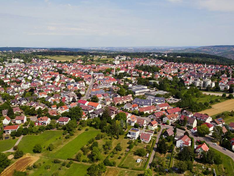 Eine Luftaufnahme zeigt eine weitläufige Wohnsiedlung mit vielen Häusern, grünen Flächen und einem leicht hügeligen Hintergrund.