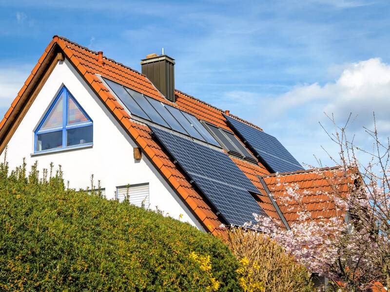 Ein Haus mit roten Dachziegeln und mehreren Solarpanels auf dem Dach, umgeben von blühenden Bäumen. /KI generiert)