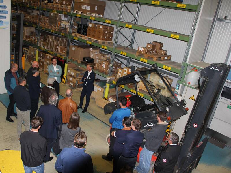 Eine Gruppe von Menschen steht in einem Lager vor einem Gabelstapler und Regalen mit Kartons und beobachtet einen Mann im Anzug. (KI generiert)