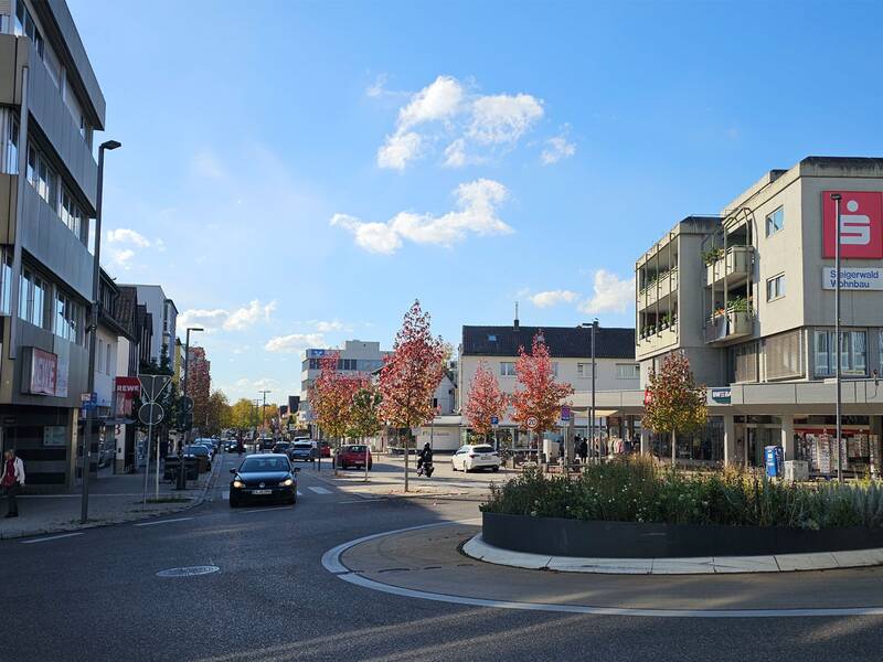 Eine städtische Straßenszene mit mehreren Autos, Gebäuden auf beiden Seiten und einigen herbstlich gefärbten Bäumen unter einem klaren blauen Himmel. (KI generiert)