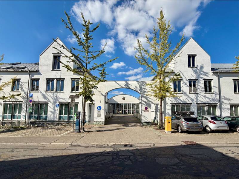 Ein modernes, weißes Gebäude mit Giebeldach und einem Torbogen in der Mitte, umgeben von jungen Bäumen, mit parkenden Autos und einem blauen Himmel im Hintergrund. (KI generiert)