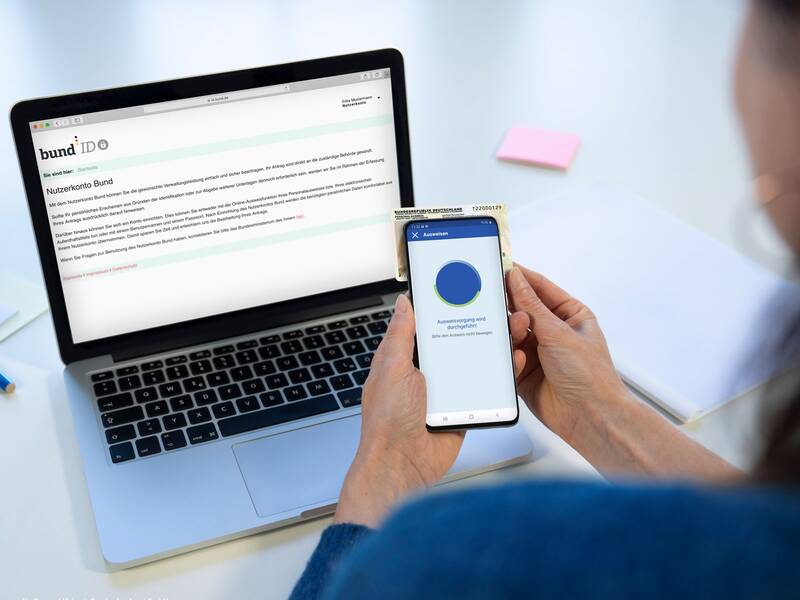 Eine Person hält ein Smartphone mit einer ID-Karte davor, während im Hintergrund ein Laptop eine Webseite mit der Überschrift "Nutzerkonto Bund" anzeigt.