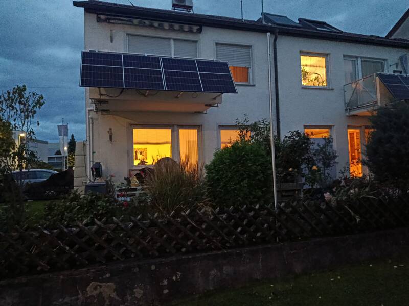 Ein zweistöckiges Haus mit Solarpanelen auf dem Dach, dessen Fenster warm beleuchtet sind, steht in einem Garten mit einem niedrigen Zaun. (KI generiert)