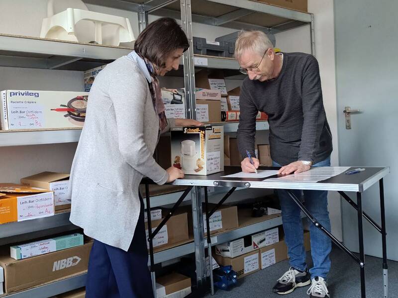 Zwei Personen stehen an einem Tisch in einem Lagerraum, auf dem Produkte in Regalen lagern, während eine Person Notizen macht. (KI generiert)