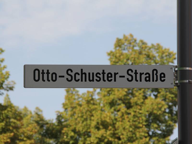 Ein Straßenschild mit der Aufschrift "Otto-Schuster-Straße" steht vor einem Hintergrund mit grünem Laub und blauem Himmel. (KI generiert)
