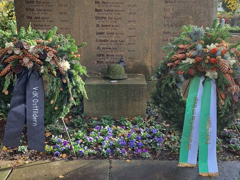 Zwei Gedenkkränze stehen vor einem Steinmonument mit eingravierten Namen, umgeben von violetten Blumen. (KI generiert)