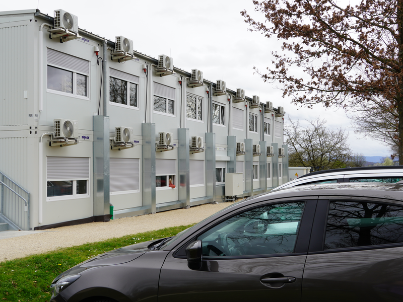Mehrere Container mit Fensterreihen und angebrachten Klimaanlagen stehen nebeneinander, im Vordergrund sind parkende Autos und ein Baum sichtbar.