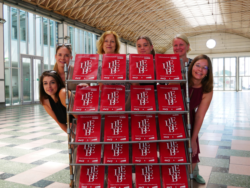 Fünf Personen posieren lächelnd um einen Zeitschriftenständer mit mehreren Ausgaben, die das Wort „LIEBE“ in großen Buchstaben zeigen, in einer Halle mit gewölbter Holzdecke und großen Fenstern.