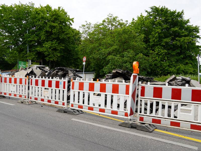 Eine Baustelle ist mit weiß-roten Absperrgittern gesichert, hinter denen aufgebrochener Asphalt zu sehen ist, umgeben von Bäumen.