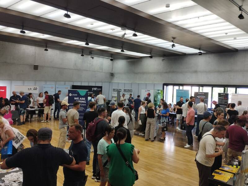In einem modernen, gut beleuchteten Raum findet eine Messe oder ein Networking-Event statt, bei dem viele Menschen um Informationsstände versammelt sind und sich unterhalten.