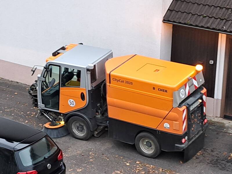 Ein orangefarbenes Kehrfahrzeug mit der Aufschrift "CityCat 2020" steht auf einem asphaltierten Hof neben einem Gebäude.