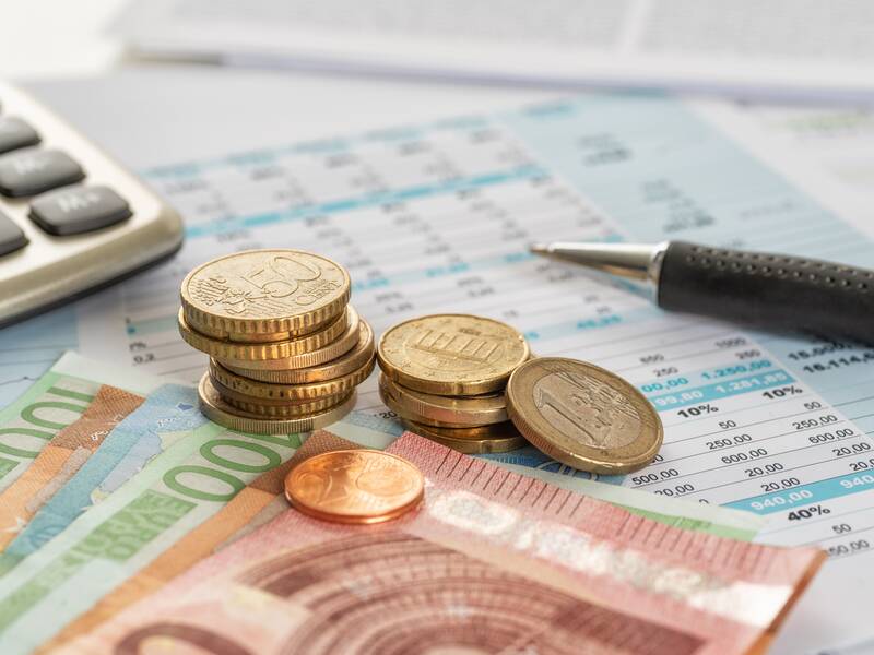 Mehrere Euro-Geldscheine und -münzen liegen auf einem Tisch neben einem Kugelschreiber und Taschenrechner auf Finanzdokumenten.