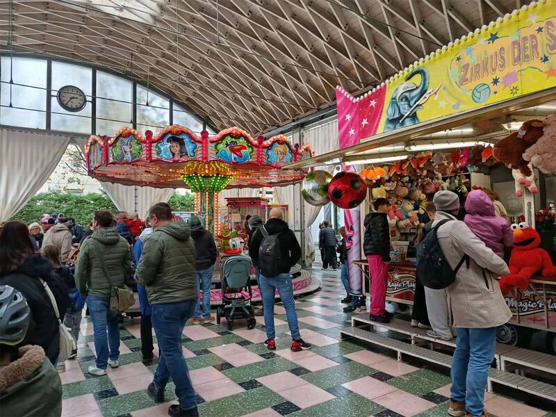 Eine bunte Karussellszene in einer überdachten Passage mit Menschen, die das Treiben beobachten, und einem Stand voller Plüschtiere. (KI generiert)