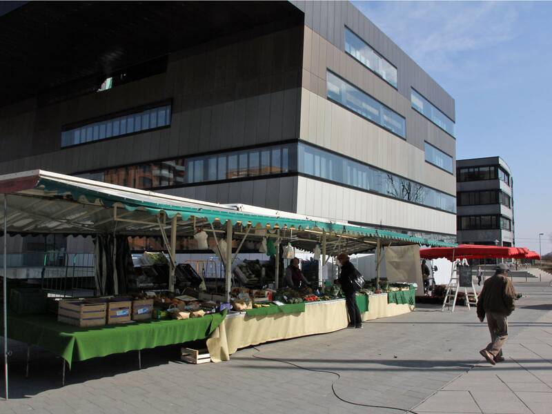 Ein Wochenmarktstand mit grünem Tuch bietet Gemüse und andere Lebensmittel an, während im Hintergrund ein modernes Gebäude zu sehen ist. (KI generiert)