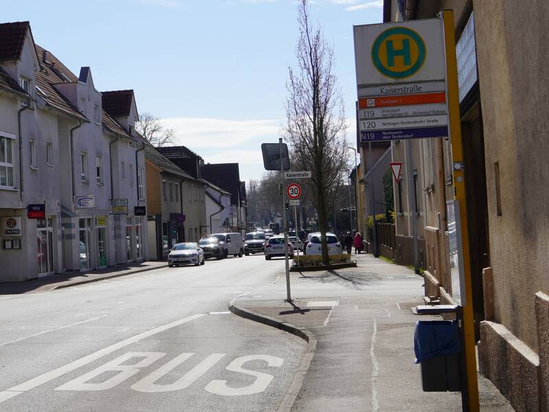 Eine Straße mit einer Bushaltestelle namens "Kaiserstraße" auf einer sonnigen Straße, entlang derer mehrere Autos und Geschäfte zu sehen sind.