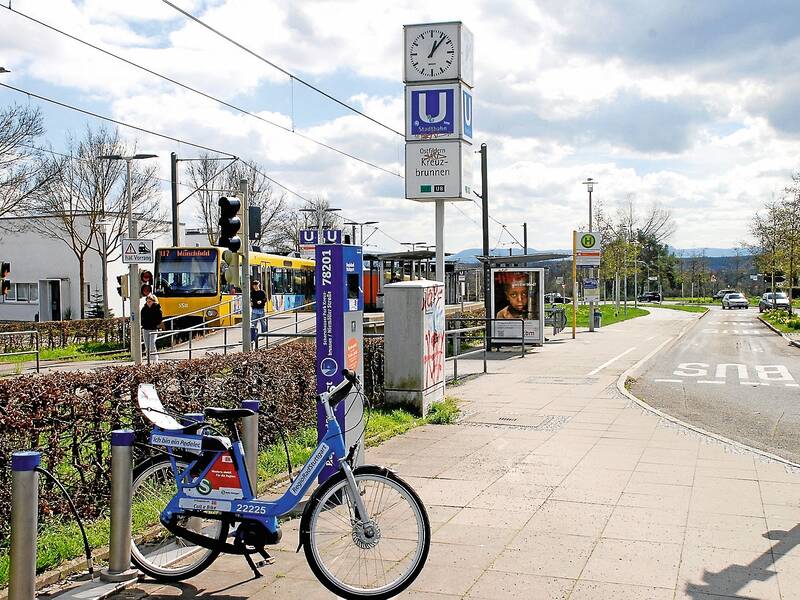 Eine belebte Straße mit einer Straßenbahnhaltestelle, einem gelben Zug, einem blauen Leihfahrrad im Vordergrund und einer Uhr über dem Stationsschild.