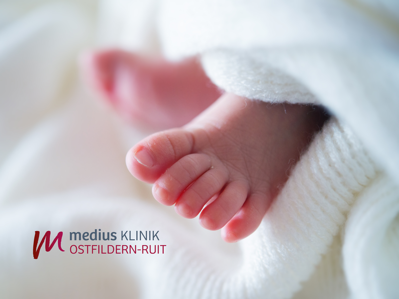 Ein kleines Baby liegt in eine weiße Decke gewickelt, und im Vordergrund sind die winzigen Zehen zu sehen; Links unten befindet sich das Logo der medius KLINIK OSTFILDERN-RUIT.
