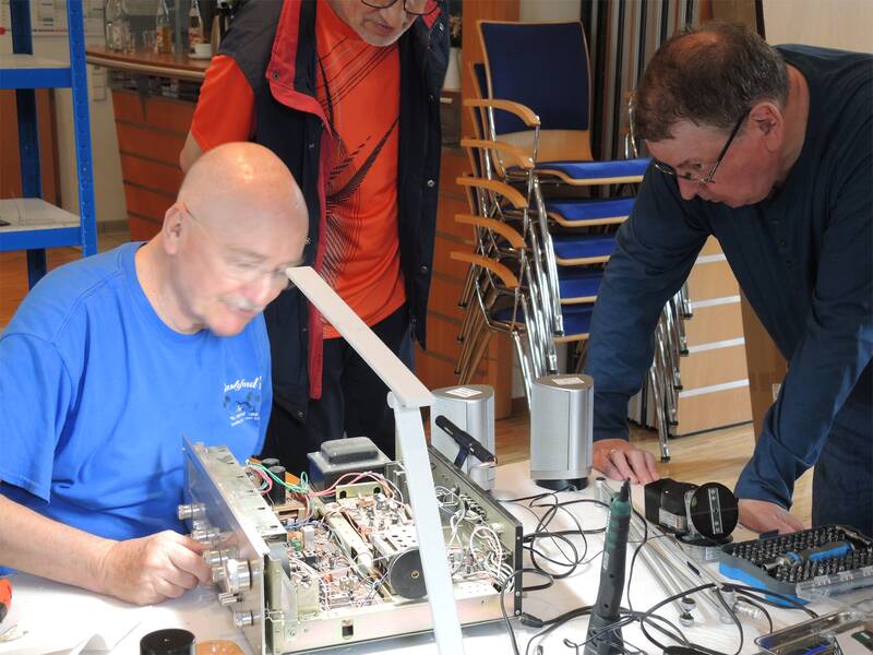 Drei Männer untersuchen gemeinsam das geöffnete Innere eines elektronischen Geräts in einem Raum mit gestapelten Stühlen im Hintergrund.