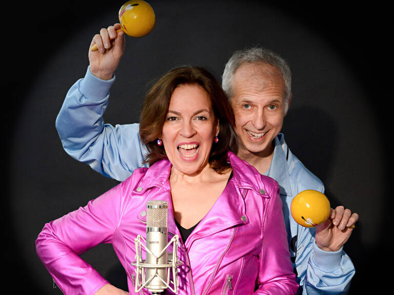 Ines Martinez und Bobbi Fischer Ein Mann und eine Frau lachen fröhlich in die Kamera; sie trägt eine leuchtend pinke Jacke, er hält gelbe Maracas und beide stehen vor einem Mikrofon.