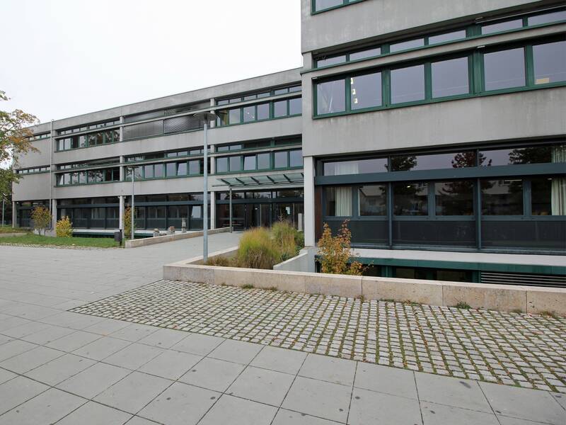 Die Realschule mit großen, rechteckigen Fenstern und einem gepflasterten Vorplatz, umgeben von ein wenig grüner Vegetation.