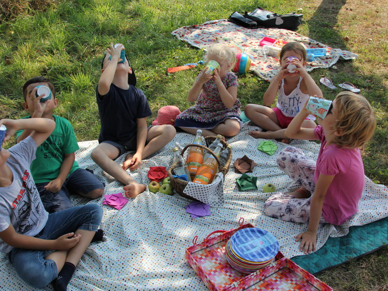 Sechs Kinder sitzen auf einer Wiese und trinken aus Bechern, während sie auf einer Picknickdecke mit Snacks und Getränken sitzen.