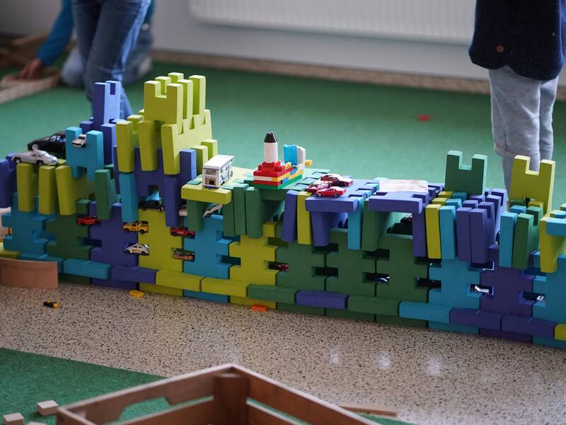 Ein Kind baut eine bunte Mauer aus großen Spielbausteinen, auf der verschiedene kleine Spielzeugautos verteilt sind.