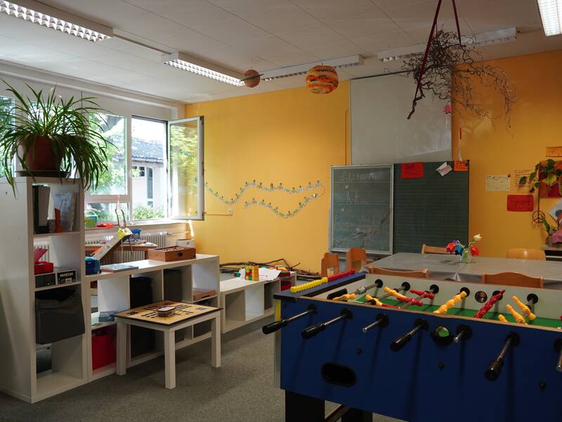 Ein bunter und einladender Betreuungsraum mit einem Tischkicker, einer Kreidetafel und Regalen voller Spiele und Material.