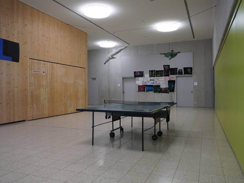 Ein leerer Raum mit Tischtennisplatte in der Mitte, einer Holzwand links, einer grünen Wand rechts und bunten Zeichnungen an der weißen Wand im Hintergrund.