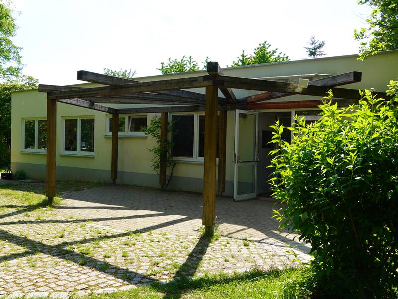 Die Schulkindbetreuung Scharnhausen ist in einem einstöckigem Gebäude mit großen Fenstern und einem offenen Eingang, umgeben von viel Grün und einem mit Holzleisten überdachten Pflasterweg, untergebracht.