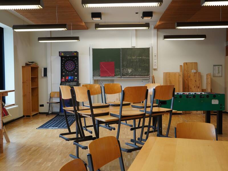 Ein Betreuungsraum mit aufeinander gestapelten Stühlen auf den Tischen, einer Dartscheibe, einer Kreidetafel mit Zeichnungen und einem Kickertisch.