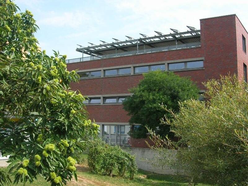 Ein modernes, dreistöckiges Backsteingebäude ist von üppiger Vegetation umgeben, mit Solarmodulen auf dem Dach.