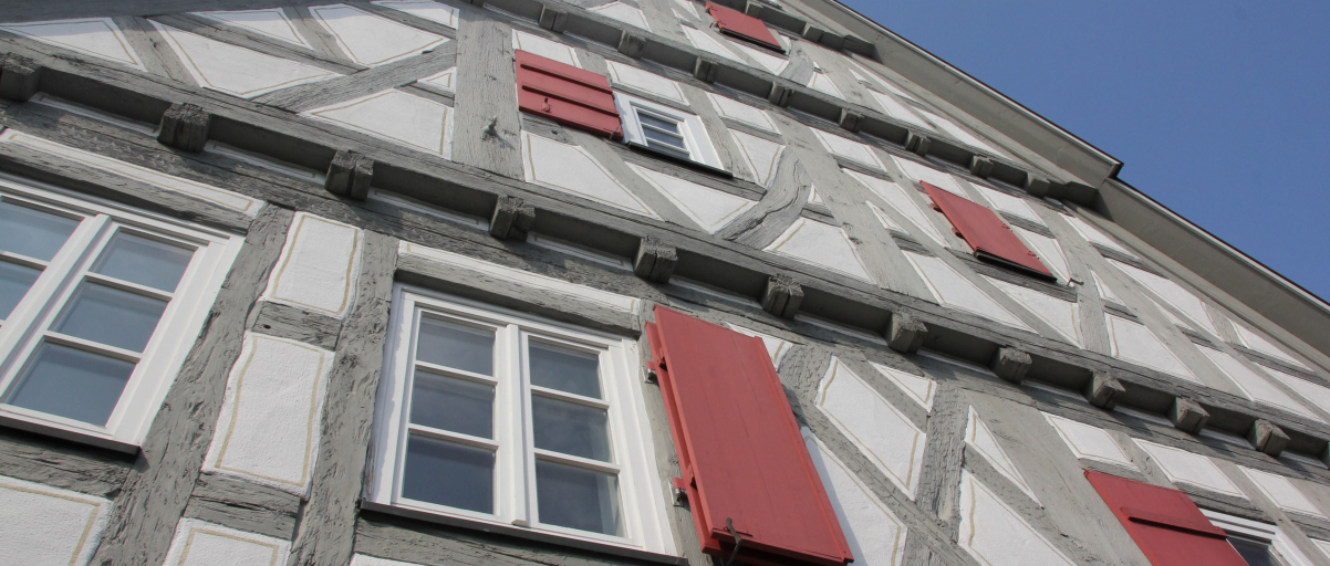 Blick an der Hauswand nach oben entlang des Nellinger Gebäudes Schopf. Das Fachwerk ist grau, die Fensterläden sind dunkelrot.