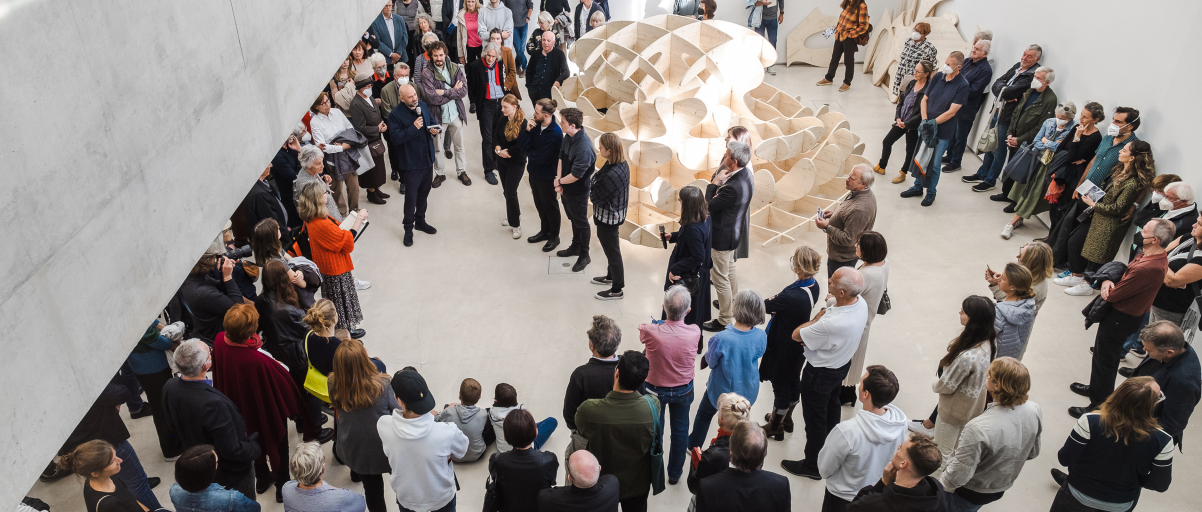 Blick von oben in die städtische Galerie. Viele Menschen stehen um ein Kunstwerk und hören den in die Ausstellung einführendenden Worten zu.