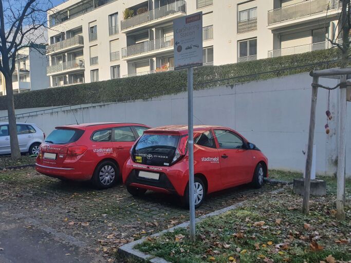 Carsharing Station: Erkennbar ist ein gepflasteter Parkplatz, auf welchem zwei rote Autos stehen. 
