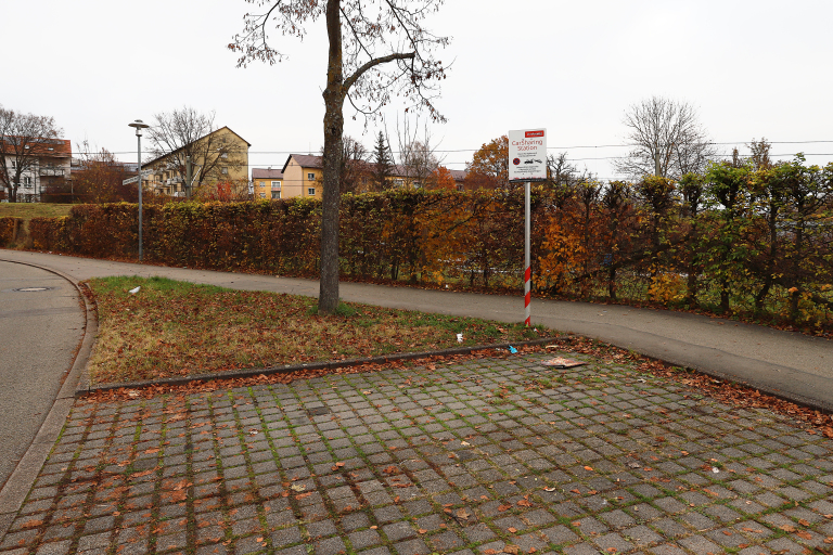 Carsharing Station: Erkennbar ist ein gepflasteter Parkplatz. Am Rand steht ein weißes Schild, welches auf einen Carsharing-Platz hinweist. 
