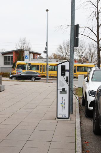 Erkennbar ist eine Ladestation für Elektromobilität. Rechts daneben sieht man zwei Autos. Im Hintergrund ist die UBahn abgebildet, die vorbeifährt.
